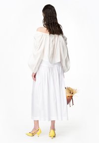Blusa bianca con spalle scoperte e maniche a palloncino, abbinata a una gonna bianca a vita alta e scarpe con tacco gialle, mentre tiene una borsa di paglia.