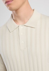 Polo beige con texture a coste verticali, patta con tre bottoni e colletto piatto. Il tessuto sembra morbido e leggero.