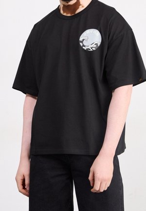 Homme portant un t-shirt noir surdimensionné avec un petit motif circulaire blanc en forme de vague sur le côté gauche de la poitrine, associé à un pantalon noir.