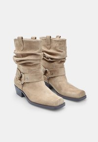 Beige velúrbőr bokacsizma laza kialakítással, széles szegéllyel, díszítő pánttal ezüst hardware-rel, és alacsony blokksarkúval.
