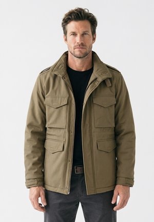 Homme portant une veste vert olive avec quatre poches avant sur une chemise noire, debout devant un fond blanc uni.