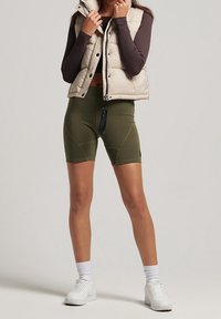 Beige vadderad väst över en brun tröja, kombinerad med olivgröna cykelshorts. Inkluderar vita sneakers och vita ankelsockor för kontrast.