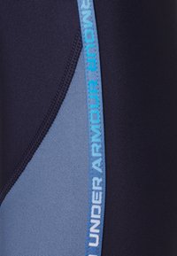 Zbliżenie na granatowy i jasnoniebieski materiał odzieżowy sportowy z jasnoniebieskim paskiem logo Under Armour wzdłuż szwu.