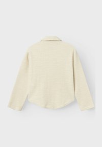 Camisa de manga larga color crema claro con un patrón tejido texturizado, que presenta un cuello puntiagudo y un dobladillo curvado, confeccionada con una tela suave.