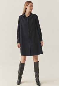 Tumes sinine nööpidega särk-kleit, millel on krae ja pikad varrukad, lõdva istuvuse ja kergelt tekstuuriga kangas. Kantakse koos kõrgete mustade saapatega.