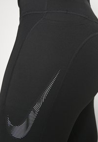 Černé sportovní legíny vyrobené z pružného materiálu s výrazným bílým logem Nike swoosh a ozdobnými švy.