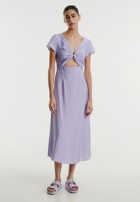 Lavendelfarbenes Kleid mit V-Ausschnitt, geknotetem Vorderteil und Ausschnitt; weicher, leichter Stoff, der bis zur Mitte der Wade fließt. Kombiniert mit passenden Sandalen.