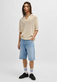Maglione corto a maniche corte di colore crema con scollatura a V, abbinato a pantaloni corti in denim blu chiaro e sneaker nere.