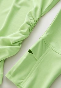 Haut vert clair avec une texture froncée sur un côté et un tissu lisse de l'autre, légèrement brillant et à la coupe ajustée.