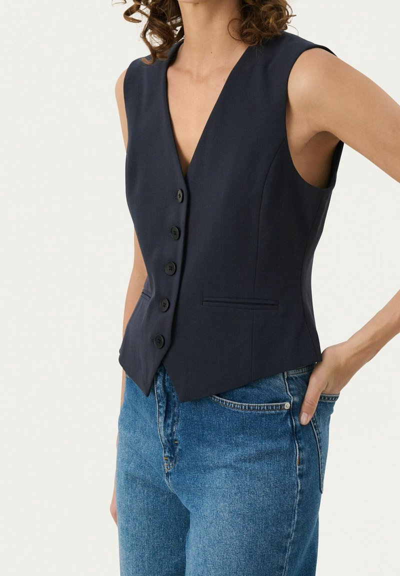 Navy-blaues ärmelloses Hemd mit V-Ausschnitt, fünf schwarzen Knöpfen, taillierter Passform und einer einzigen Vordertasche. Getragen über blauen Jeans.