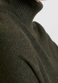 Ciemnozielony golf wykonany z teksturowanej dzianiny. Posiada składany wysoki kołnierz i opuszczone szwy na ramionach.