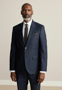 Un costume à carreaux bleu foncé avec une fermeture à un bouton, des revers crantés et un mouchoir de poche. Une chemise blanche et une cravate texturée complètent le look.