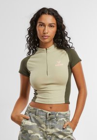 Kortermet cropped topp i lysegrønn med mørkegrønne detaljer. Har høyt krage og frontglidelås, med rosa logo-detaljer.