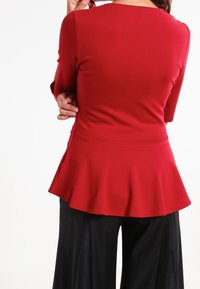 Kvinde iført en rød peplum-top med ærmer til albuen og sorte brede bukser, vist fra ryggen mod en hvid baggrund.