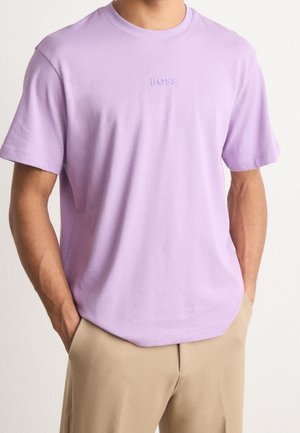 Mand iført lyse lilla kortærmet T-shirt med "BOSS" logo og beige bukser, hænderne i lommerne, foran en ensartet lys baggrund.