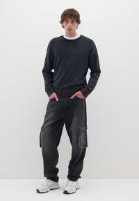 Maglione lavorato a maglia grigio scuro con colletto a coste, abbinato a pantaloni cargo neri larghi con una grande tasca laterale e scarpe da ginnastica bianche.