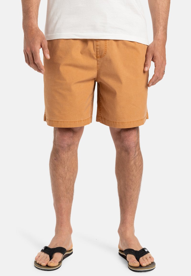 Pantalones cortos de un tejido naranja-marrón cálido, con un corte recto y dobladillo cosido. Combinados con chanclas negras. Diseño minimalista.