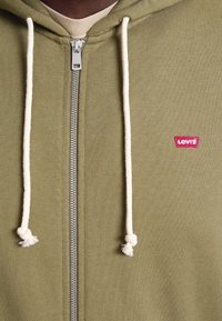 Primer plano de una sudadera con cierre en color verde olivo de Levi's, con cordones blancos y un pequeño logo rojo de Levi's en el pecho.