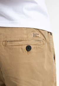 Pantaloni khaki realizzati in un tessuto testurizzato, con una tasca posteriore dotata di un bottone nero e un'etichetta con la scritta "Petrol Ind" sulla vita.