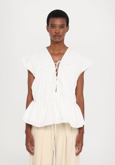 White blouse with cap sleeves, gathered design, deep V-neck, adjustable drawstrings, and a peplum hem. Smooth fabric, minimalistic style.