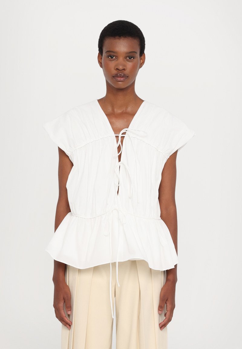 White blouse with cap sleeves, gathered design, deep V-neck, adjustable drawstrings, and a peplum hem. Smooth fabric, minimalistic style.