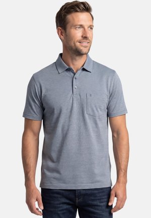 Mężczyzna w jasno niebieskiej, krótkoterminowej koszulce polo z kieszonką na piersi i ciemnych dżinsach, lekko zwracający głowę na bok, uśmiechający się.