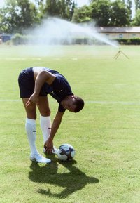 Un joueur de football Puma en maillot bleu et en short blanc se penche pour faire ses lacets près d'un ballon de football noir et blanc sur un terrain de gazon.