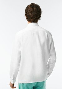 Camicia bianca a maniche lunghe realizzata in tessuto leggero, con colletto standard e polsini con bottone, vista da dietro. Texture liscia.