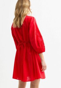 Rode linnen jurk met pofmouwen, een gathered taille met strikdetail, en bloemengalerie op de mouwen.