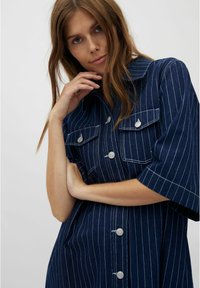 Denimkleid in Dunkelblau mit weißen vertikalen Streifen. Merkmale sind kurze Ärmel, zwei Brusttaschen und silberne Knöpfe auf der Vorderseite.