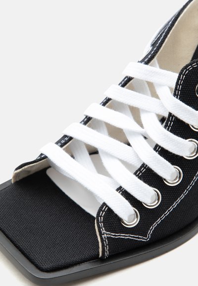 Chaussure en toile noire avec lacets blancs, œillets en métal et coutures blanches, gros plan sur la zone des orteils et des lacets.