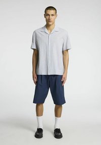 Světle modrá pruhovaná košile s krátkým rukávem a knoflíkovým zapínáním, spárovaná s tmavě modrými šortkami po kolena. Model nosí černé boty a bílé ponožky.