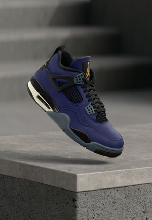 Chaussure de sport violette et noire avec des panneaux en mesh et un logo jaune, en suspension au-dessus d'un bloc de béton avec des escaliers en arrière-plan.