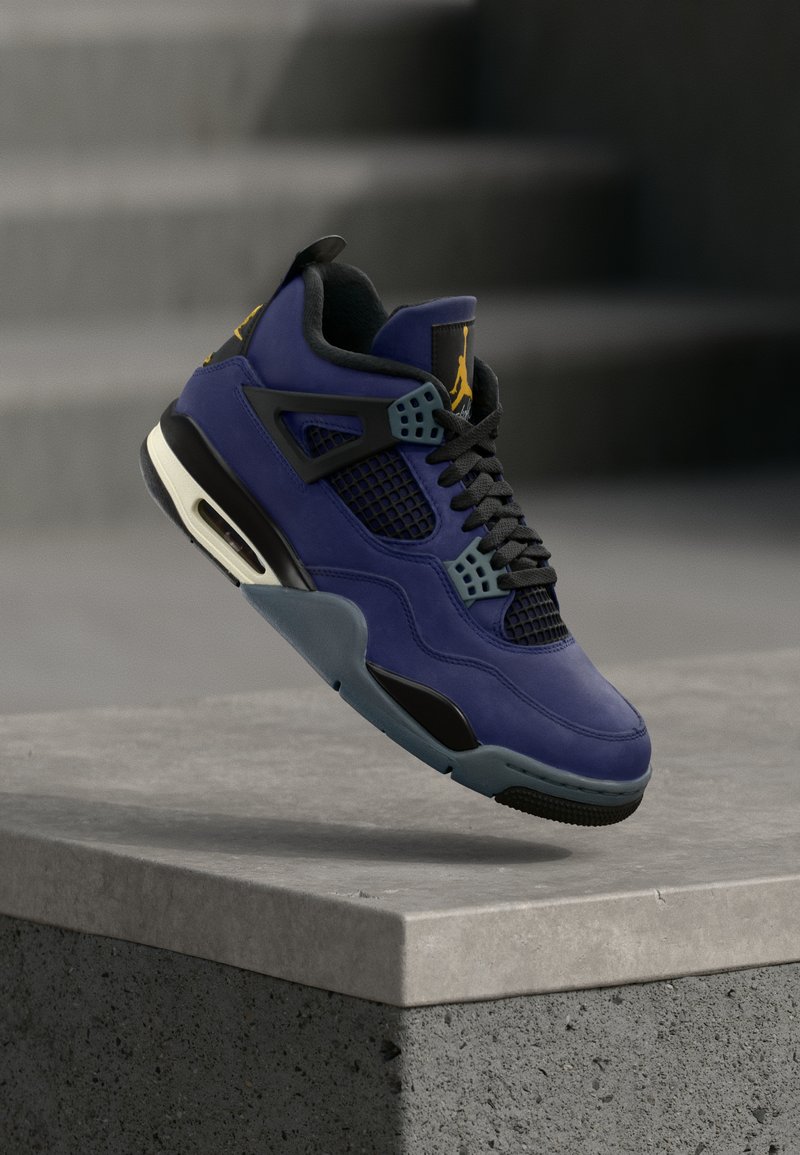 Μωβ και μαύρο αθλητικό παπούτσι με πάνελ από πλέγμα και κίτρινο λογότυπο, αιωρούμενο πάνω από μια σκυρόδετη βάση με σκάλες στο βάθος.