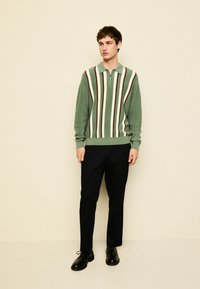 Groen-wit gestreepte trui met rits en geribbelde textuur, gecombineerd met zwarte broek en zwarte schoenen, staand tegen een neutrale achtergrond.