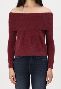 Pull en Bourgogne à épaules dénudées en tissu doux et texturé, avec des manches longues et une coupe courte, associé à un jean en denim foncé.