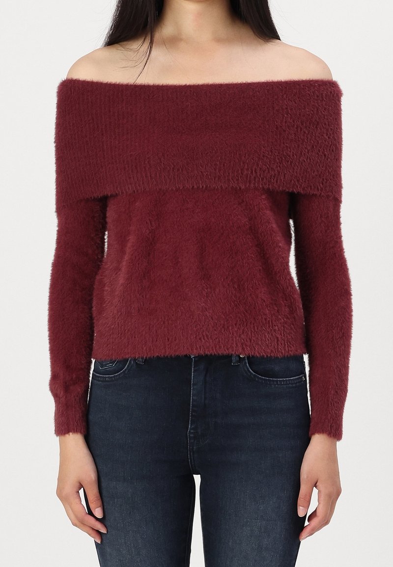 Pull en Bourgogne à épaules dénudées en tissu doux et texturé, avec des manches longues et une coupe courte, associé à un jean en denim foncé.