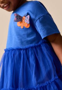 Enfant portant une robe bleue avec jupon en tulle et un patch papillon en tissu sur la poitrine, souriant et légèrement tourné sur le côté.