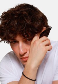 Bracelet en perles noires au poignet, porté avec un T-shirt blanc. Présente des cheveux noirs bouclés et une peau lisse dans un gros plan, avec une expression sérieuse.