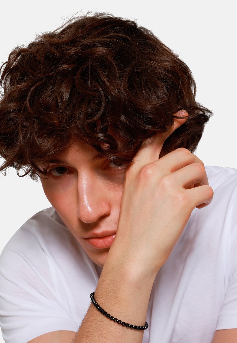 Bracelet en perles noires au poignet, porté avec un T-shirt blanc. Présente des cheveux noirs bouclés et une peau lisse dans un gros plan, avec une expression sérieuse.