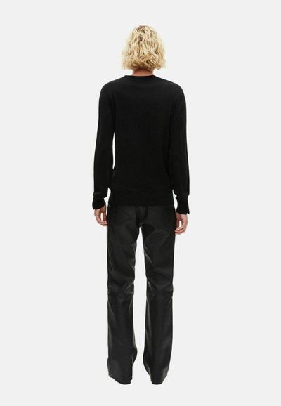 Persona con capelli biondi corti e ricci che sta di spalle, indossando una maglietta nera attillata a maniche lunghe e pantaloni di pelle nera.