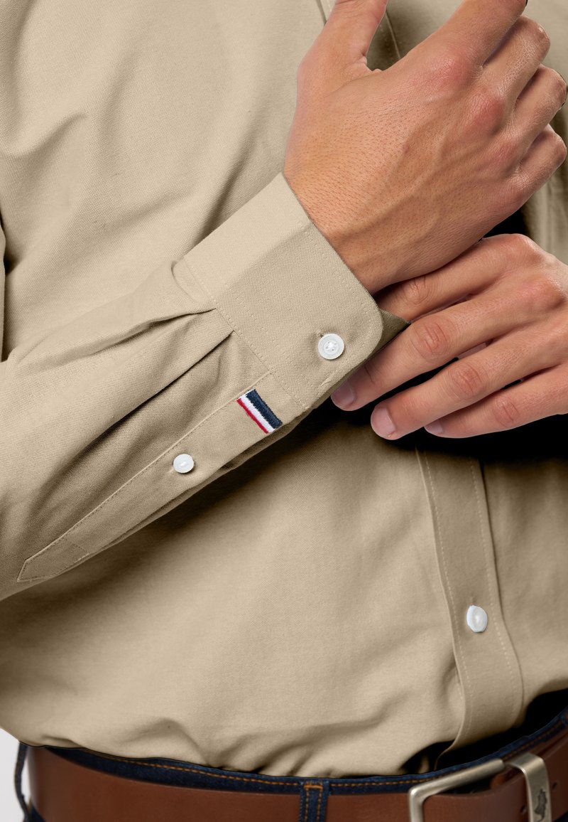 Beiges Hemd aus Baumwolle mit Knöpfen, klassischem Kragen, weißen Knöpfen und einem kleinen blauen, roten und weißen Streifenakzent an der Manschette.