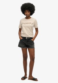 Beige bomulds t-shirt med "Superdry" logo, parret med sorte denim shorts med frynset kant. Sorte slidesandaler fuldender looket.