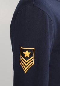 Sudadera azul marino con manga ajustada que presenta un emblema bordado en oro de dos chevrones y una estrella sobre un fondo negro.