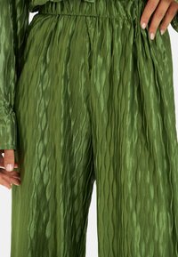 Pantalones verdes plisados de tela brillante con patrones de ondas verticales, con una cinturilla elástica fruncida y una textura suave.