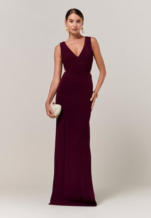 Ženska stoji v brezrokavni globoko vijolični večerni obleki z V-izrezom, drži majhno belo torbico, nosi zlate uhane in zapestnico.