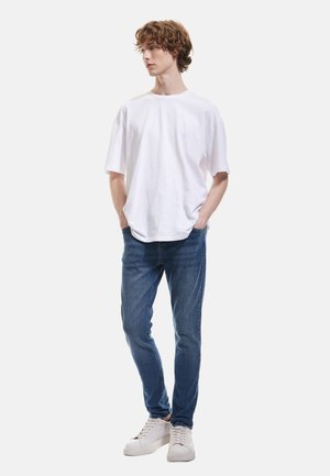 Camiseta de algodón blanca con un corte holgado, combinada con jeans de mezclilla azules. El modelo también lleva zapatillas blancas, de pie frente a un fondo simple.