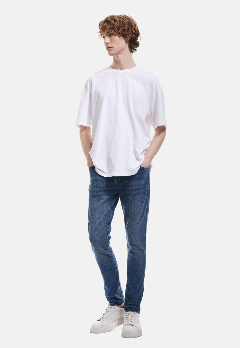 Camiseta de algodón blanca con un corte holgado, combinada con jeans de mezclilla azules. El modelo también lleva zapatillas blancas, de pie frente a un fondo simple.