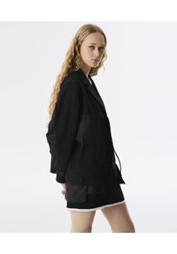 Chaqueta negra con rayas, que presenta un corte relajado, grandes bolsillos y cordones. Llevada sobre una falda corta negra. Textura suave.