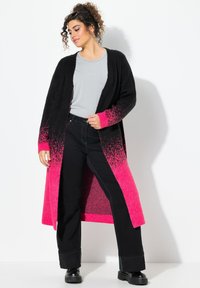 Cardigan long noir et rose dégradé avec une coupe ample, tricot texturé, devant ouvert et manches longues. Porté par-dessus un haut gris et un pantalon noir.